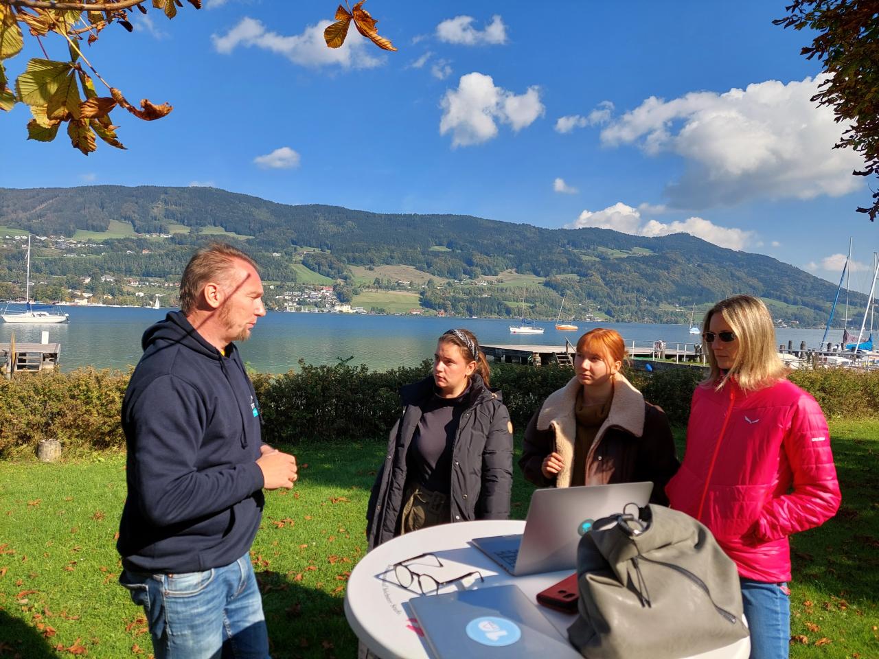 Am Ufer eines Sees stehen 4 Personen rund um einen Stehtisch