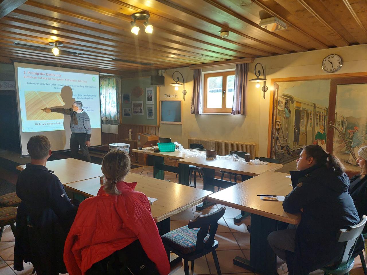 In einem Saal hält jemand einen Vortrag vor einer kleinen Gruppe von Menschen. 