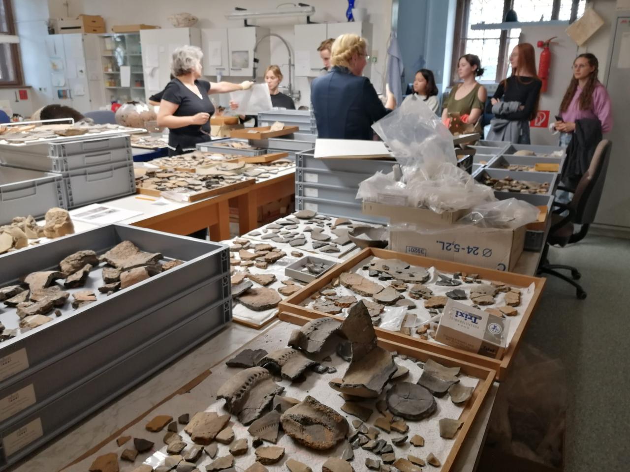 Man sieht viele Keramikfragmente auf Tischen in der Restaurationswerkstätte, die von einer Personengruppe betrachtet werden