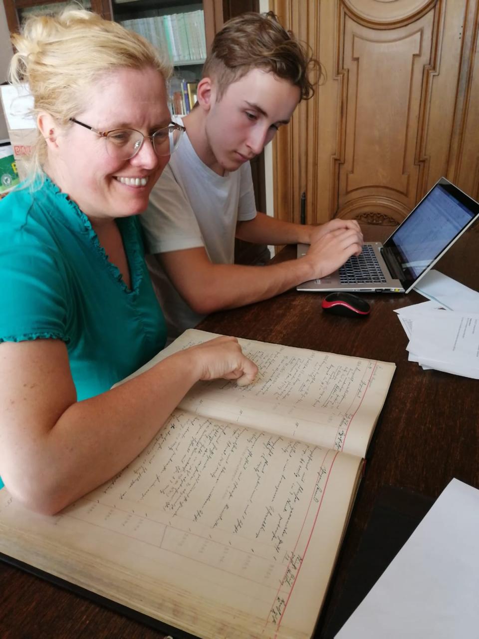 2 Personen vor einem Laptop und einem Inventarbuch an einem Schreibtisch sitzend