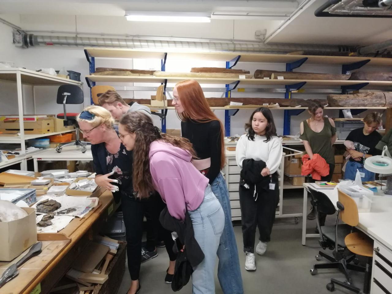 Eine Personengruppe bewegt sich in einem Archiv zwischen Regalen mit Fundstücken aus Holz und Textil