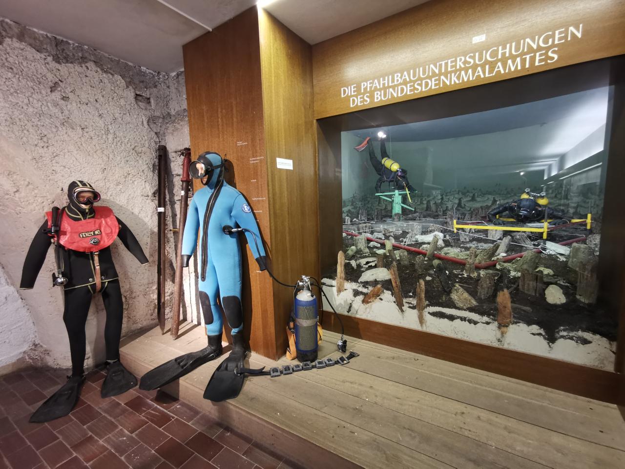 2 Taucheranzüge vor einem lebensgroßen Diorama mit Taucher:Innen
