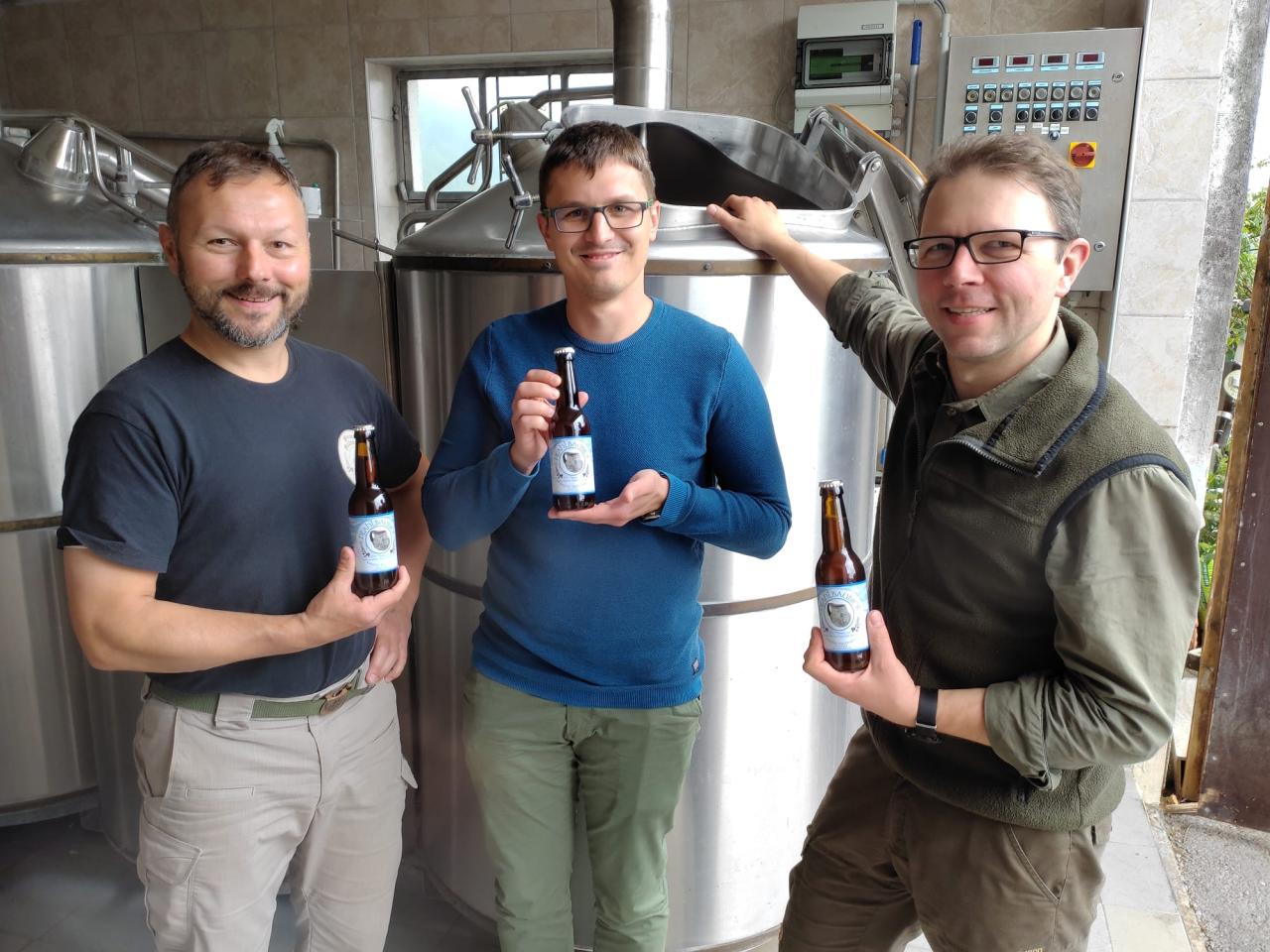 3 Männer vor einem Braukessel, jeder hält eine Flasche Bier in Händen