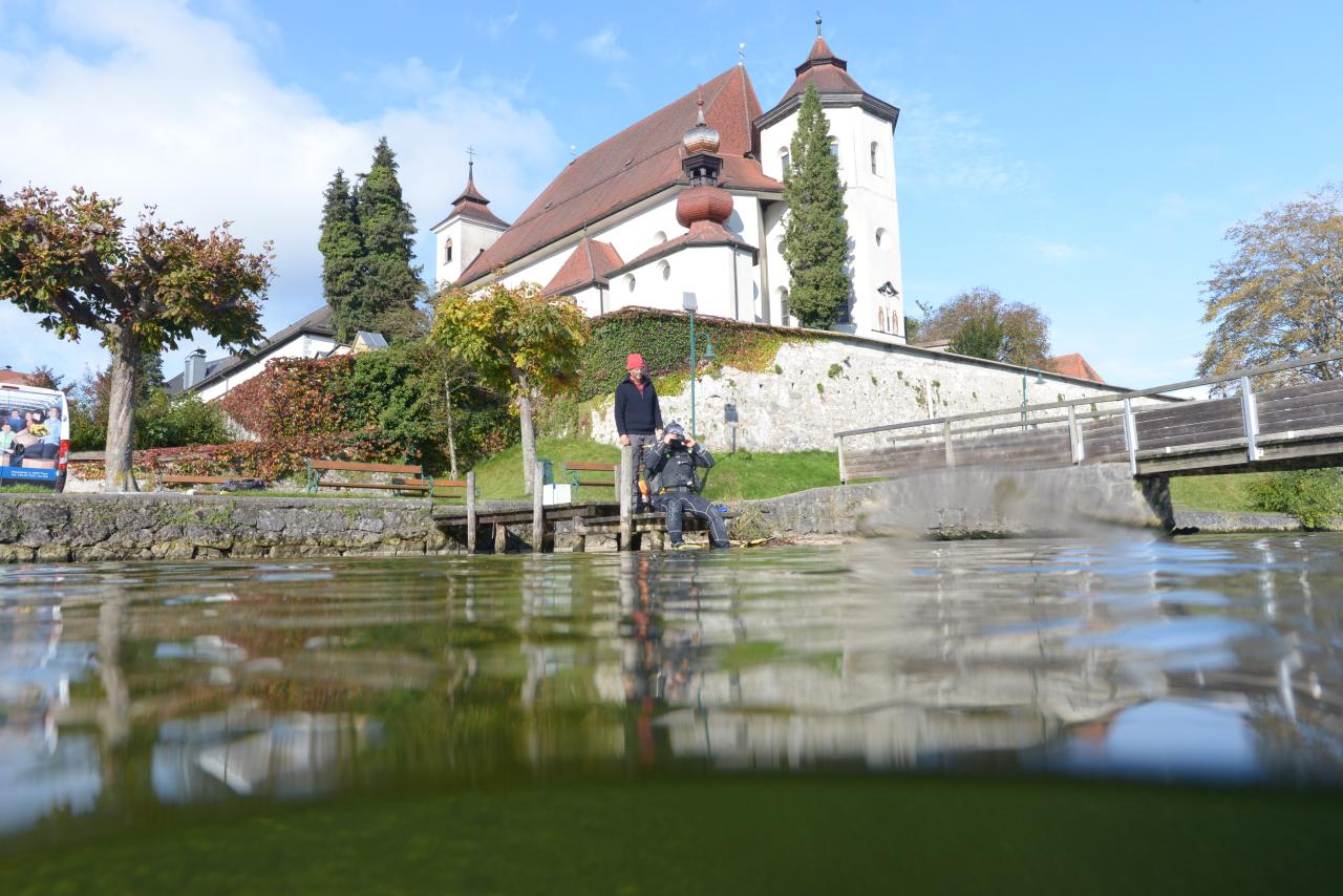 Man sieht 2 Personen am Ufer des Traunsees vor dem Kloster Traunkirchen