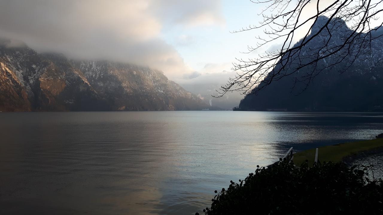 Ein See in grau- und schwarz-Schattierungen, mit den nebelverhangenen Bergen im Hintergrund