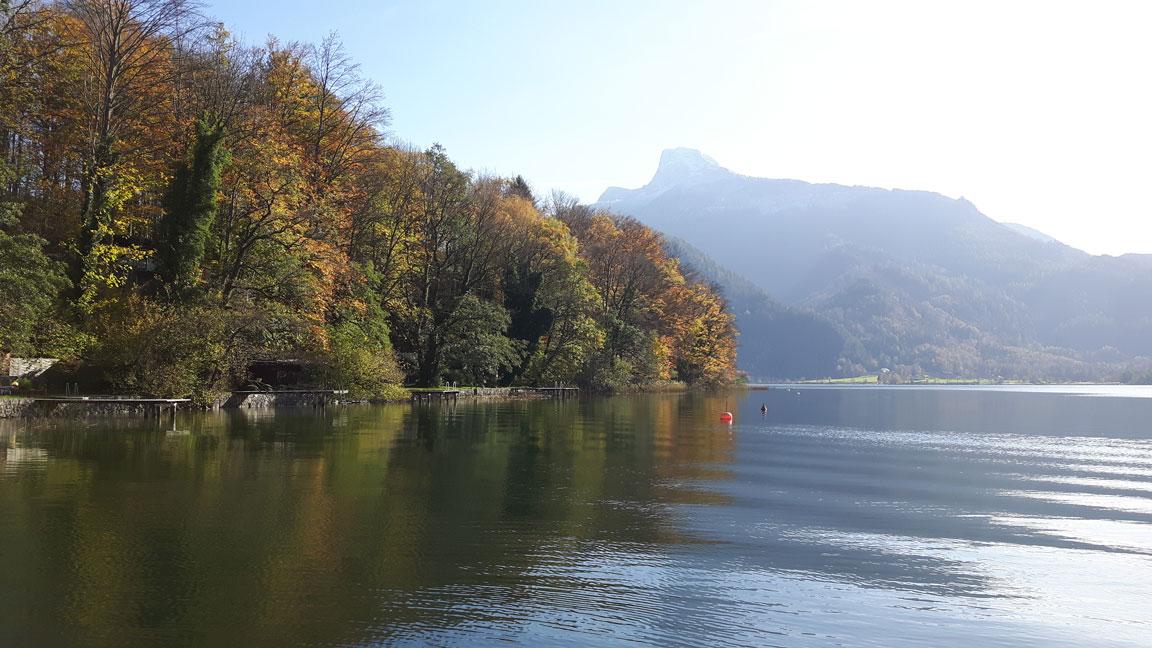 Ein See, im Hintergrund bewaldete Berge mit Herbstlaub
