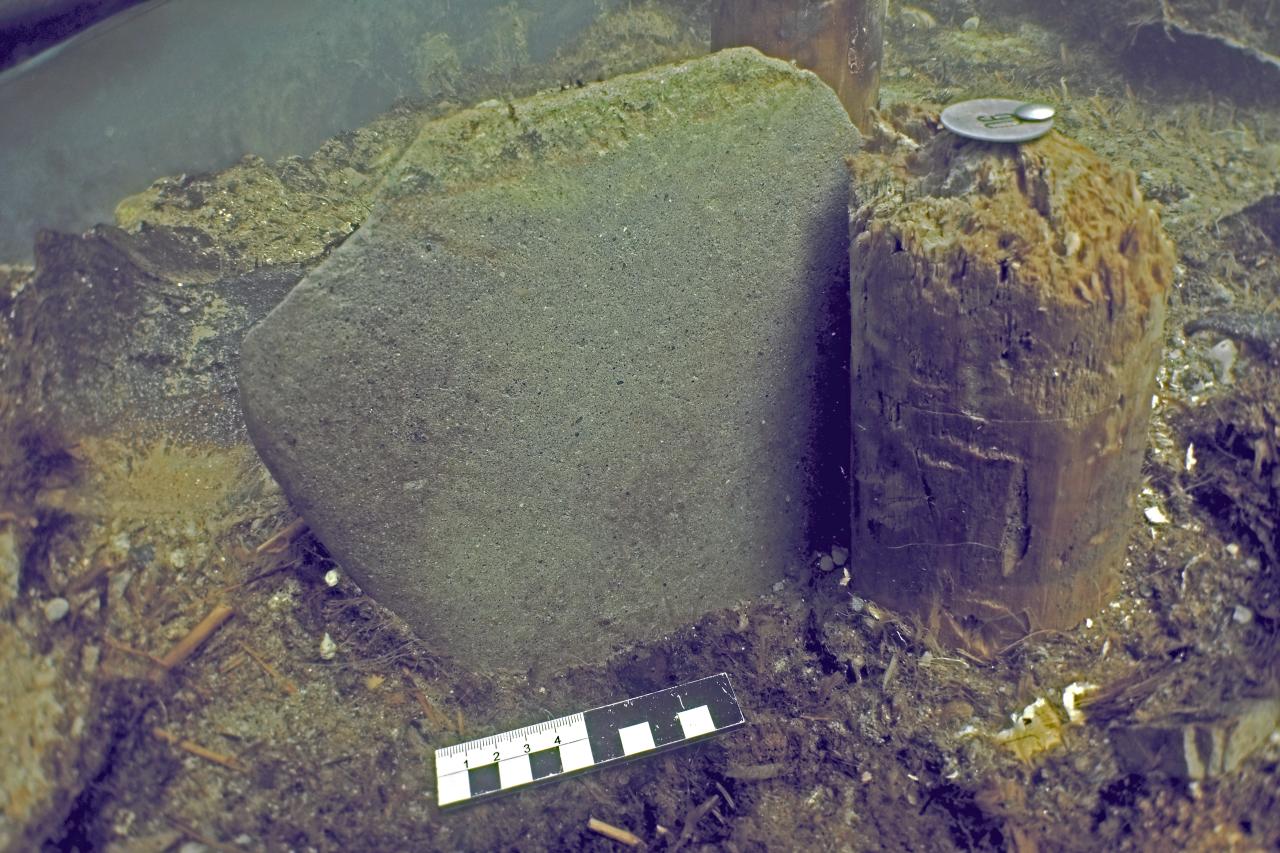 Ein Keilstein und ein Stück Holz unter Wasser, das mit einem Nagel markiert wurde. Ein Maßstab liegt davor