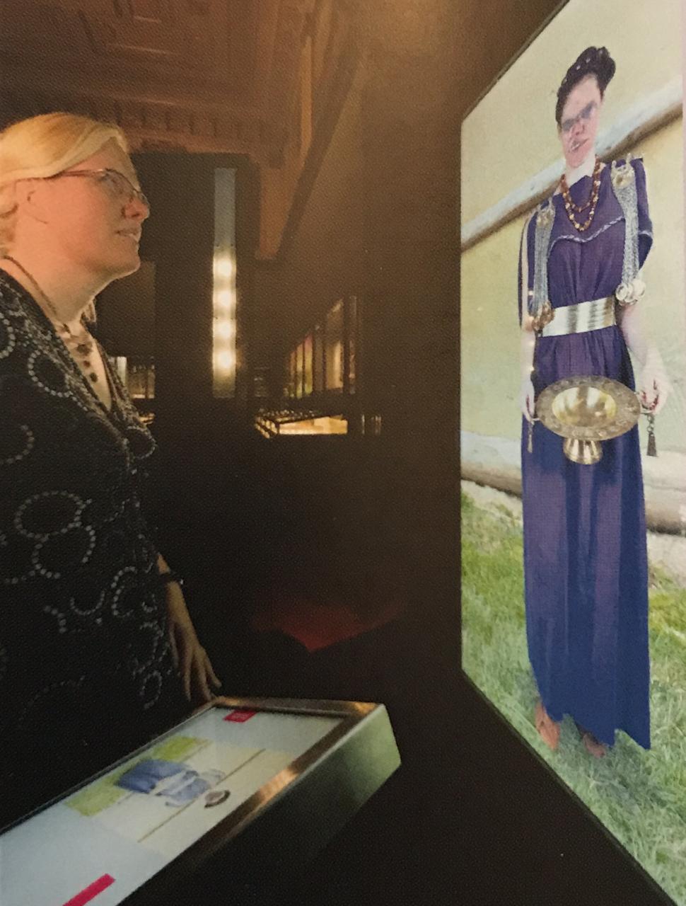 Eine blonde Frau betrachtet eine Wandprojektion einer Frau in "Hallstatt-Tracht"
