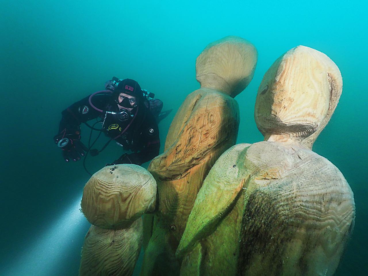 3 Holzfiguren unter Wasser, im Hintergrund ein Taucher