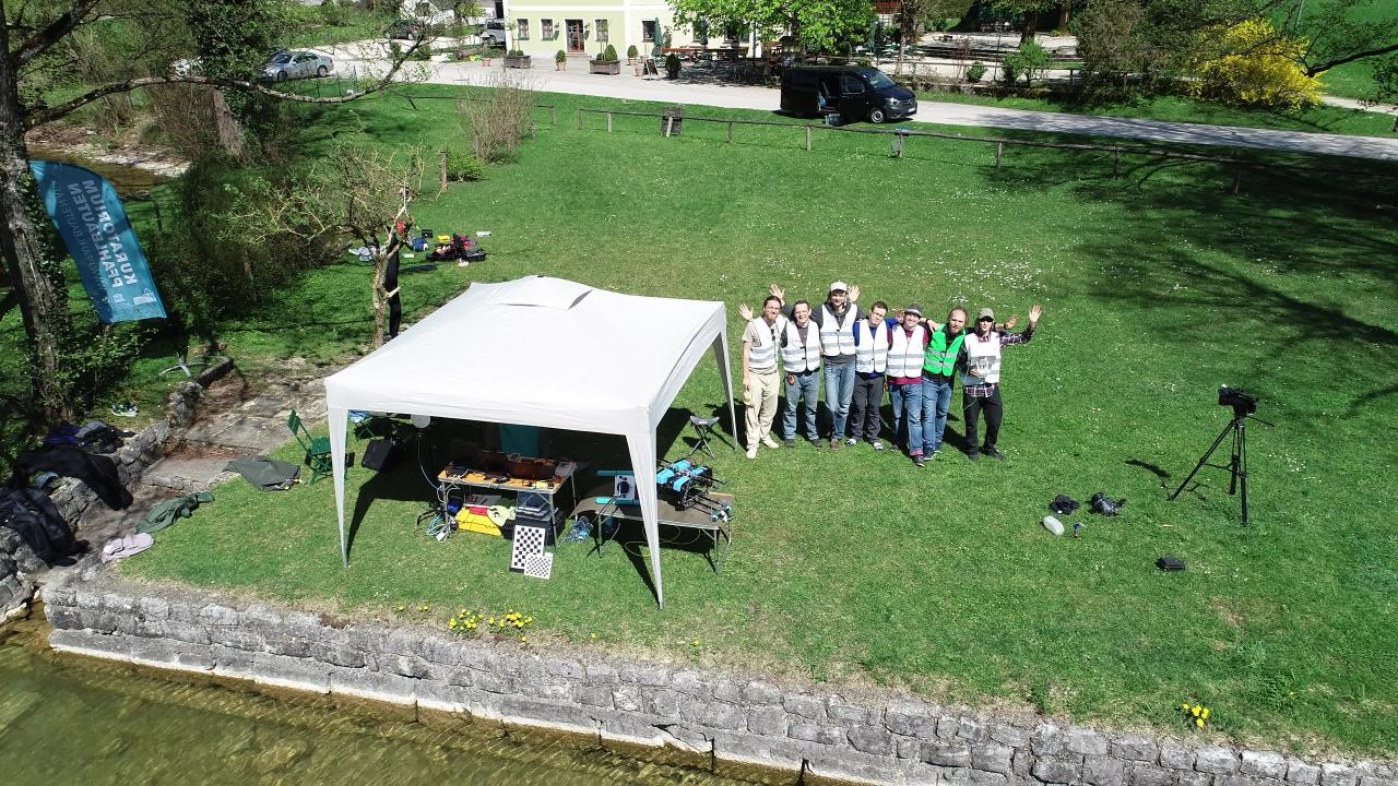 Eine Gruppe von Menschen steht am Seeufer neben einem weißen Pavillon und winkt in Richtung der Kamera