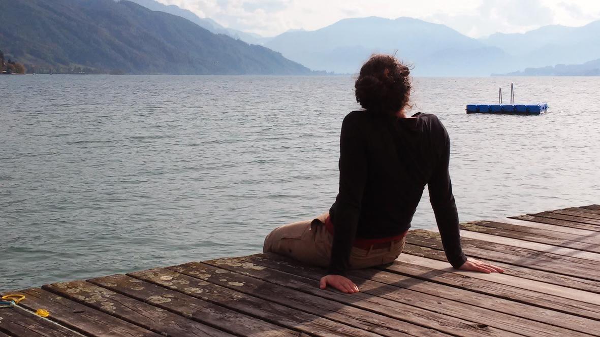 Eine Frau sitzt uns mit dem Rücken zugewandt auf einem Steg mit Blick auf den See