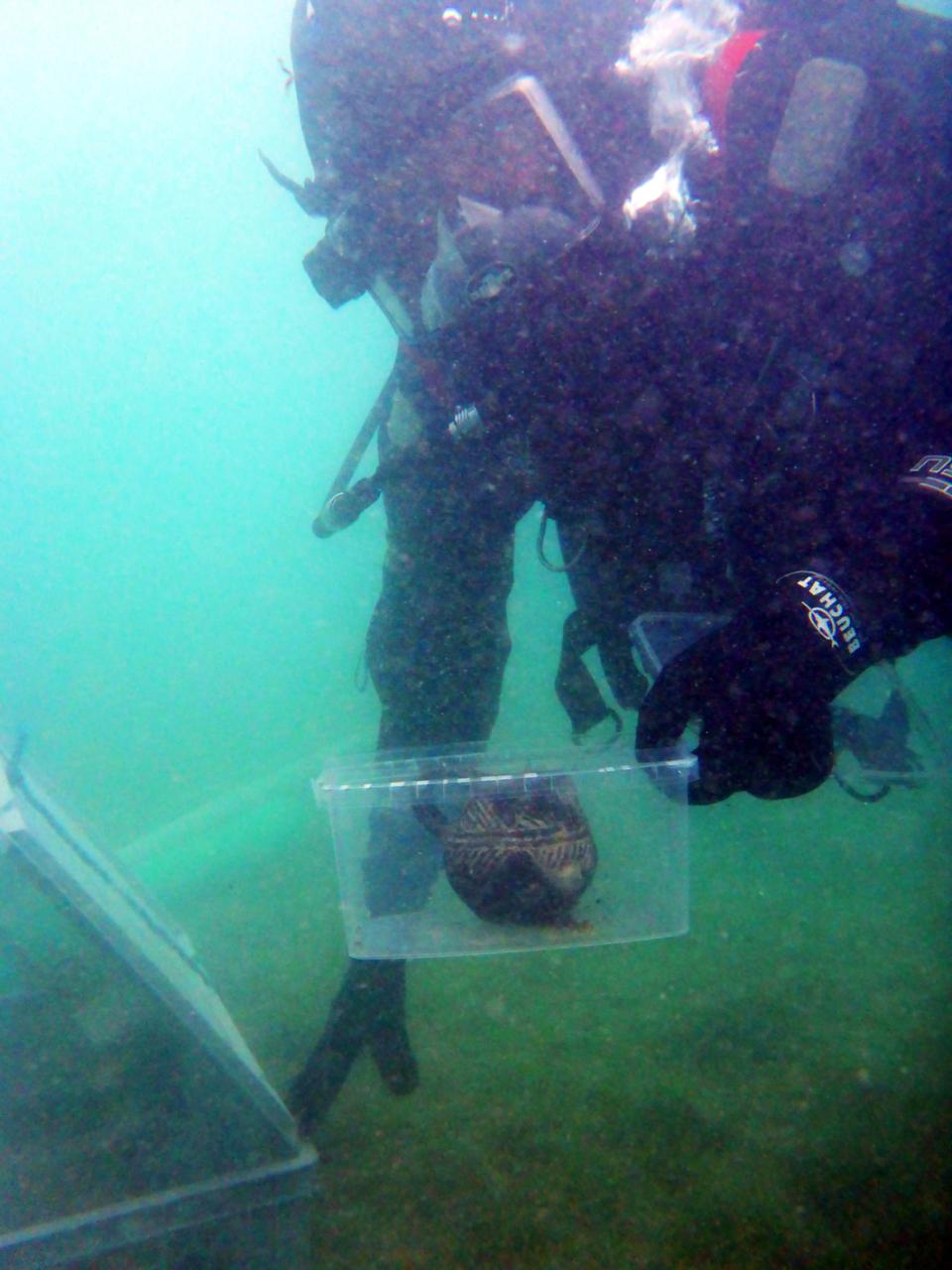 Ein Taucher unter Wasser hält ein Fundsackerl mit dem geborgenen Mondseekrug Richtung Kamera