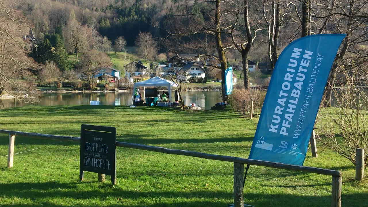 Die Fahne des Kuratorium Pfahlbauten auf einer Wiese vor dem See, im Hintergrund ein weißer Pavillon