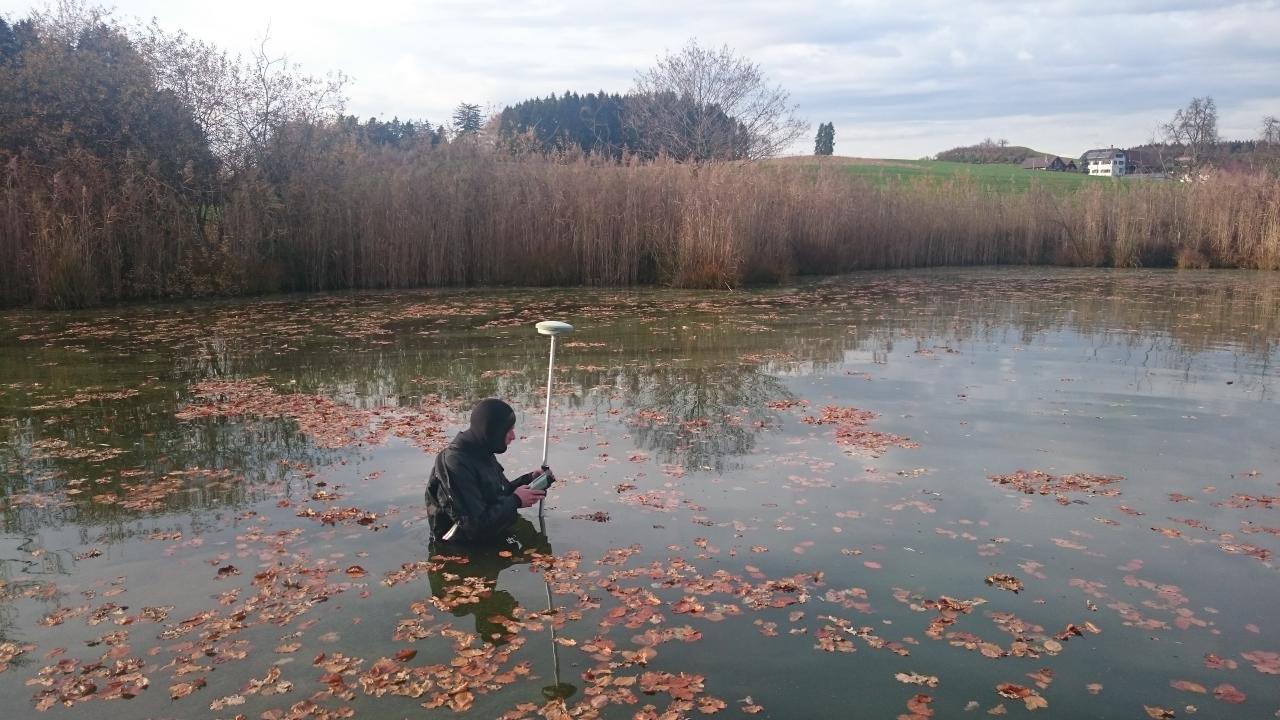 Ein Taucher steht bei Vermessungsarbeiten bis zur Hüfte im Wasser und hält ein Prisma hoch