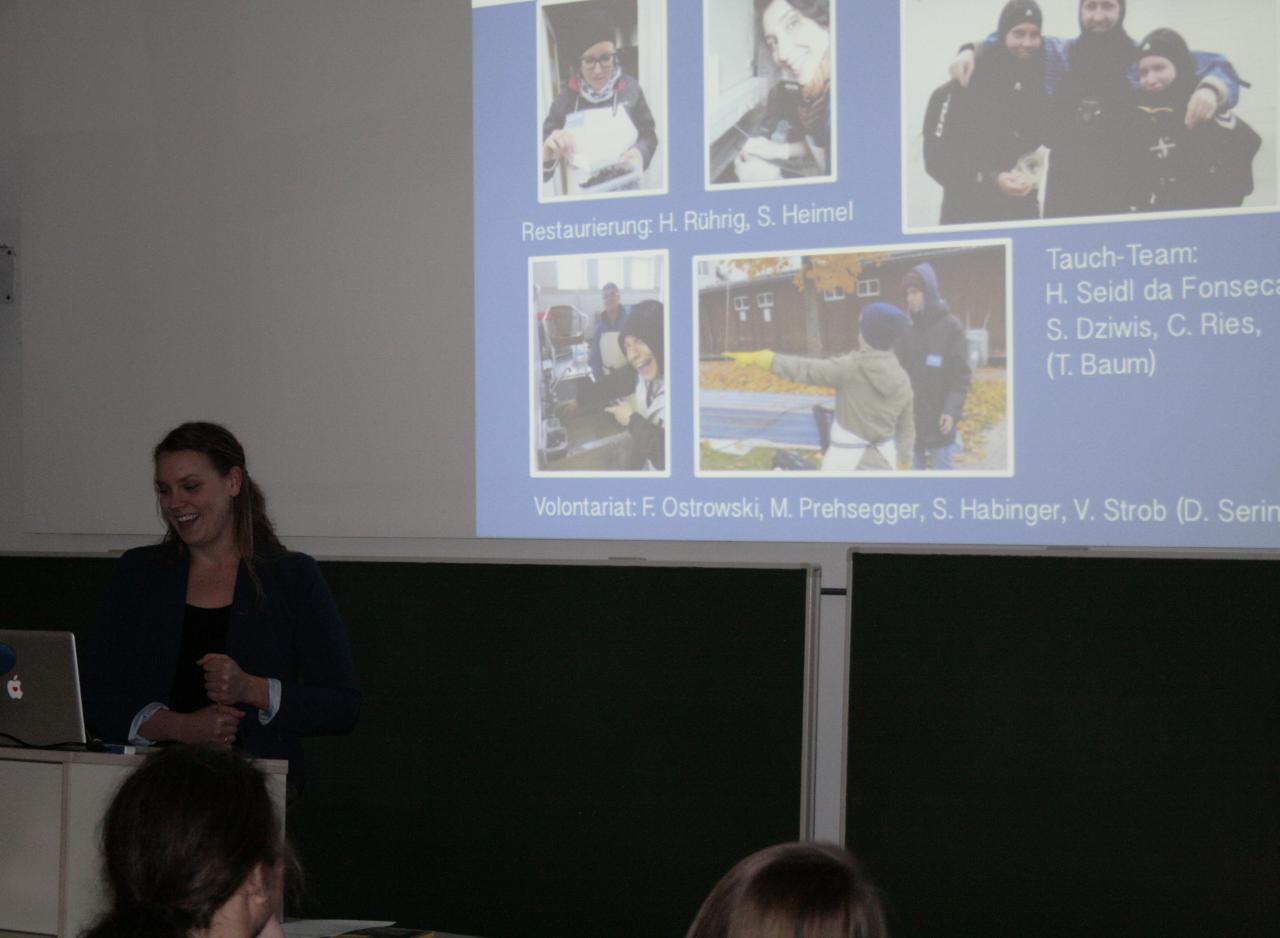 Helena Seidl da Fonseca am Rednerpult vor der Leinwand mit Bildern von der Unterwasser Ausgrabung in Seewalchen