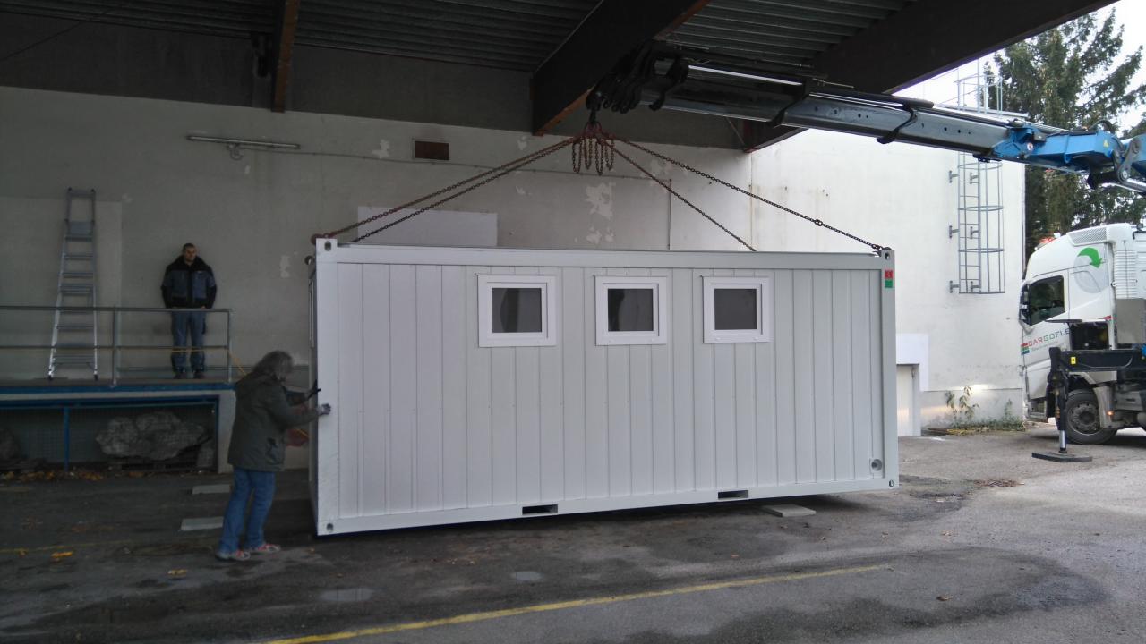 2 Arbeiter und ein Kranwagen heben den weißen Container an seinen Platz