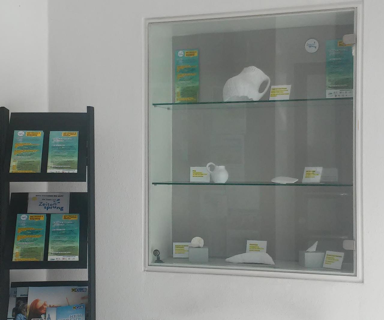 Eine Vitrine in einer weißen Wand, daneben ein Ständer mit Foldern