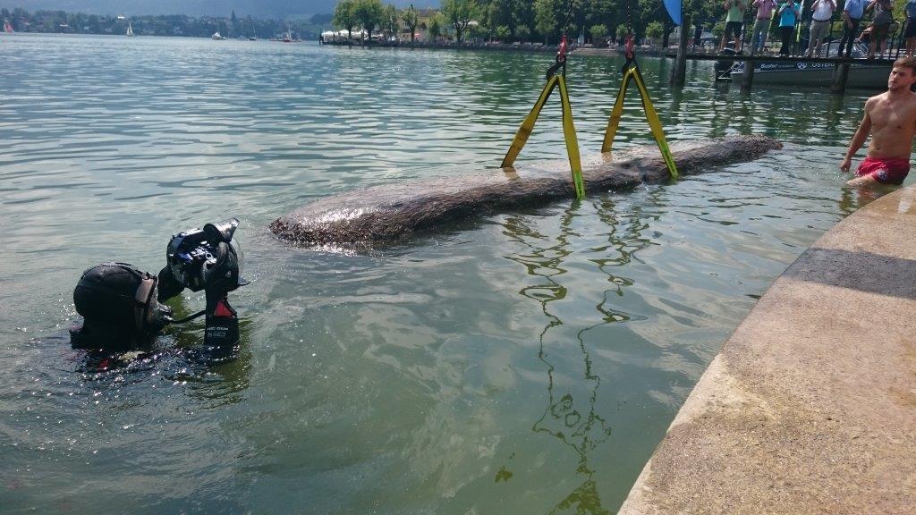 Ein Taucher fotografiert den Einbaum, der (an Gurten hängend) gerade an die Oberfläche gehoben wird