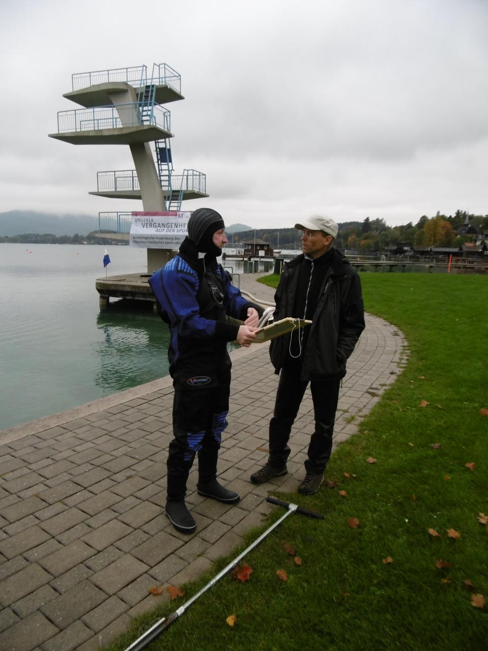 2 Männer stehen am Ufer des Strandbades Seewalchen, mit dem Sprungturm im Hintergrund