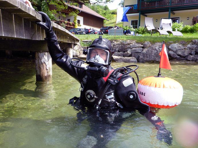 Ein Taucher mit einer Tauchboje hält sich, halb im Wasser, an einem Steg fest