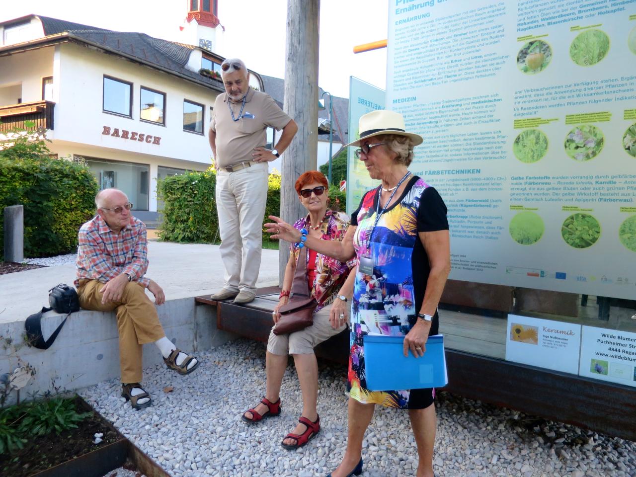 Vor einem Info-Plakat im Steinzeitgarten erklärt Helga interessierten Besucher:Innen etwas über die Anlage