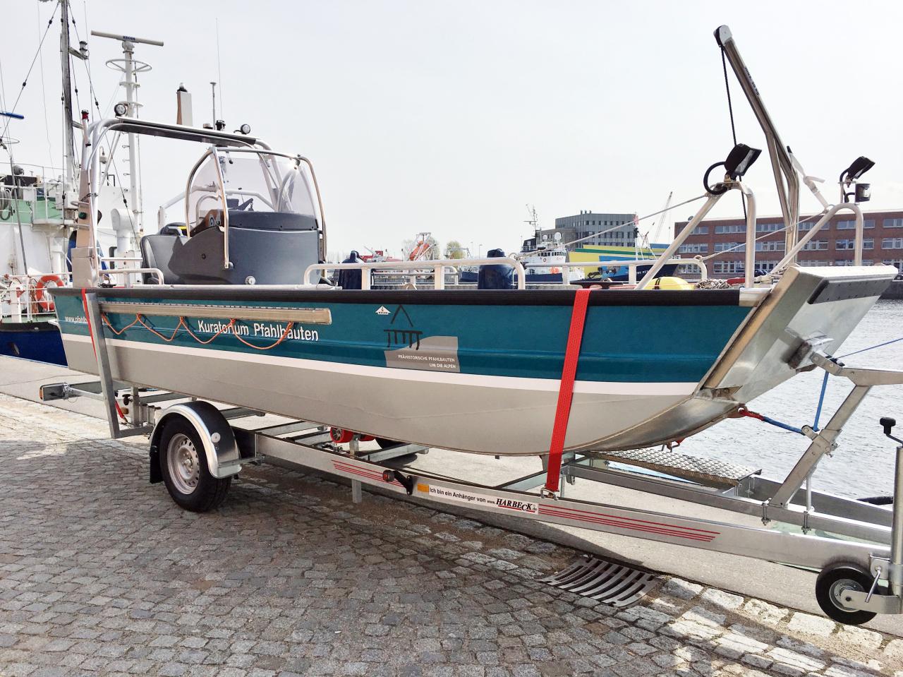 Das Forschungsboot auf dem Anhänger fixiert. Man sieht die seitliche Aufschrift "Kuratorium Pfahlbauten" und das Logo. Das Boot ist blau und silber