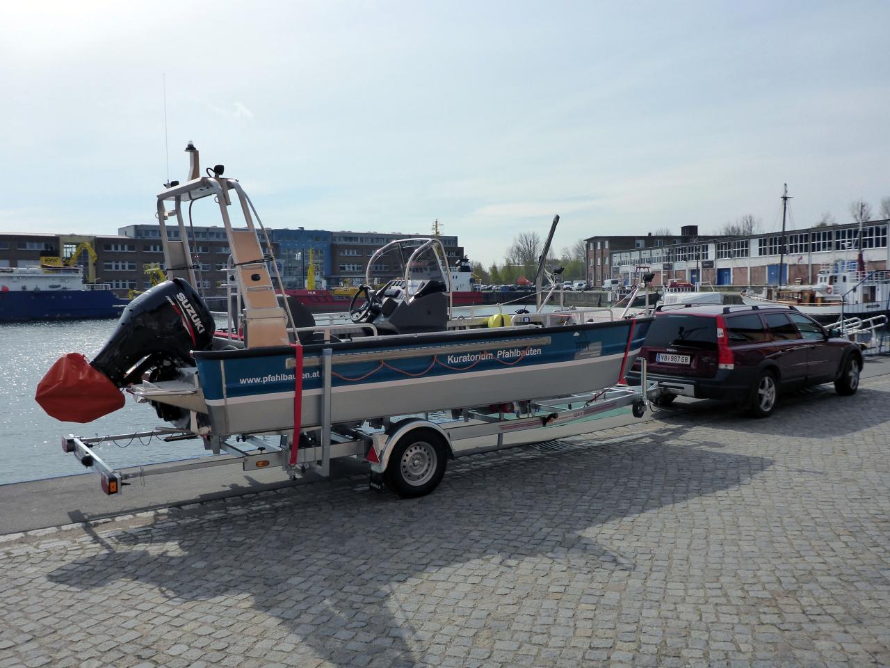 Das Forschungsboot auf dem Anhänger fixiert. Man sieht die seitliche Aufschrift "Kuratorium Pfahlbauten" und das Logo. Das Boot ist blau und silber