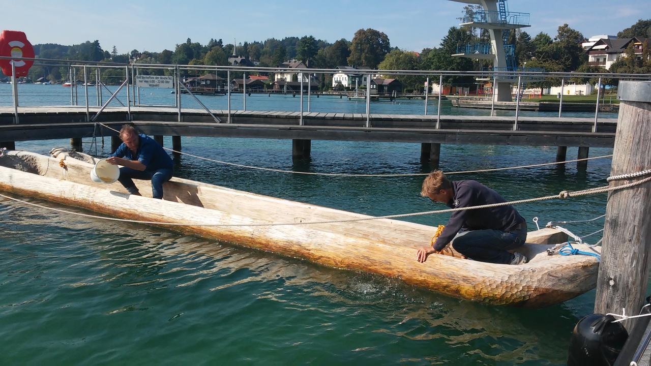 2 Personen auf einem Einbaum, der noch im Hafen vertäut liegt