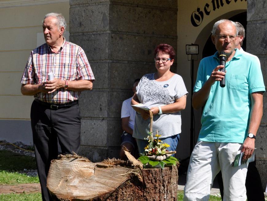 Ein Moderator steht mit 2 weiteren Personen vor einem Holzstamm