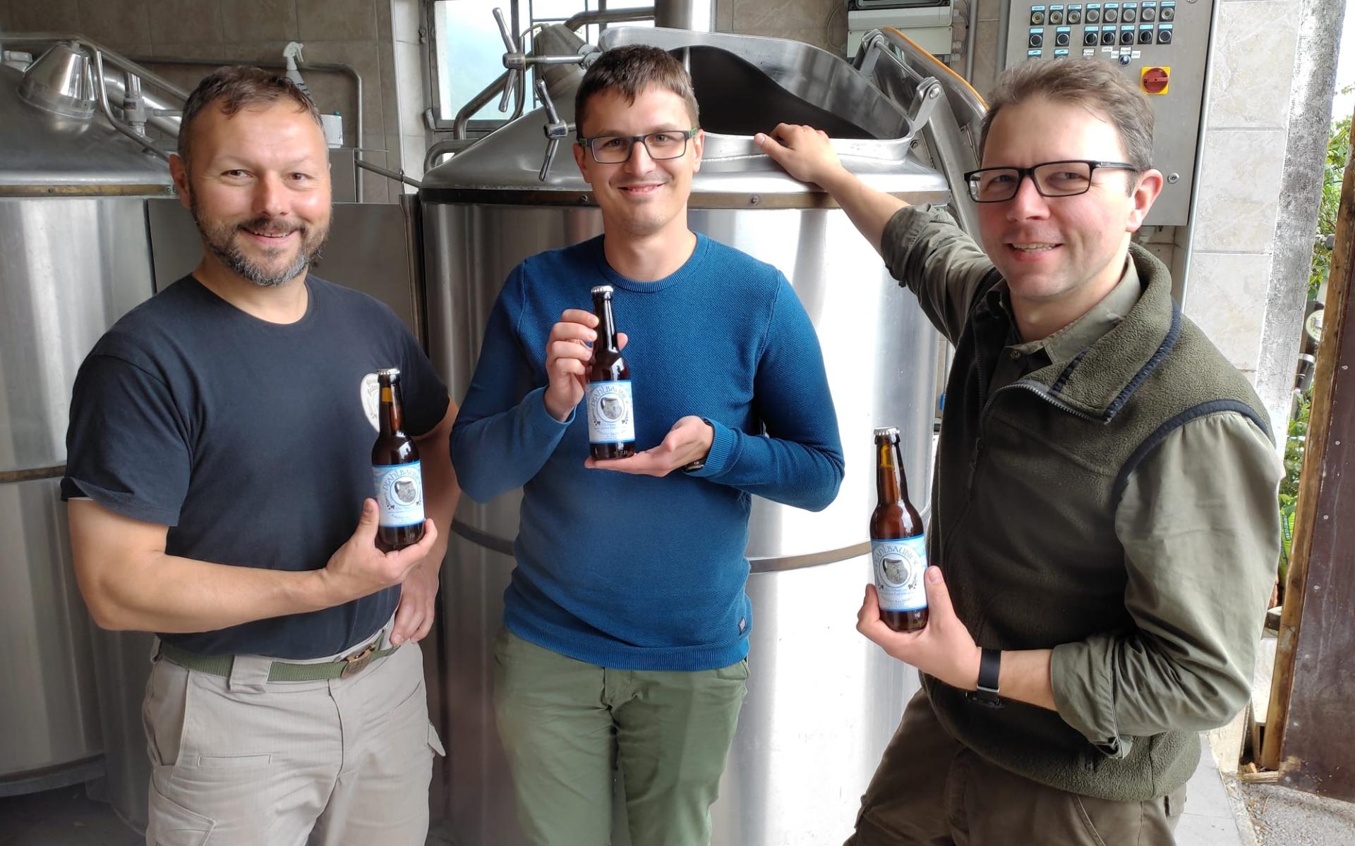 3 Männer vor einem Braukessel, jeder hält eine Flasche Bier in Händen
