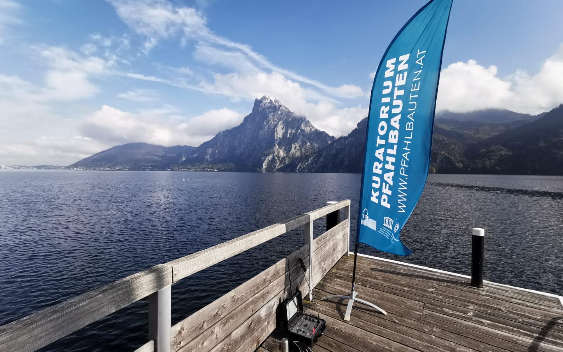 Eine blaue Beachflag des "Kuratiorium Pfahlbauten" auf einem Steg vor dem Traunsee