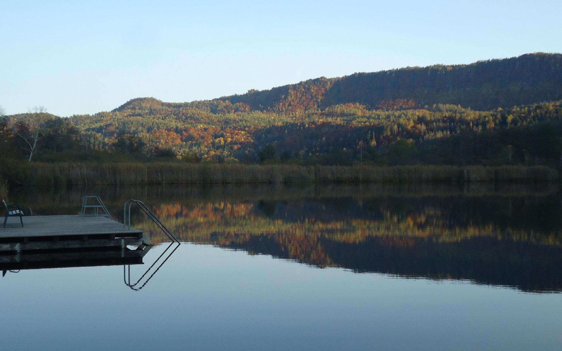 Ein See mit einem Steg, im Hintergrund ein bewaldeter Hügel