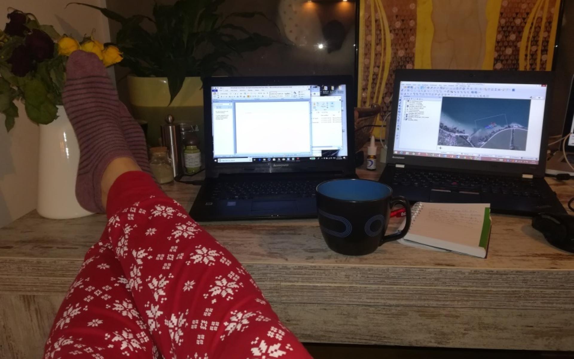 Man sieht Beine in einer roten Pyjama-Hose, die auf einem Schreibtisch liegen. Daneben eine Kaffeetasse und 2 Laptops