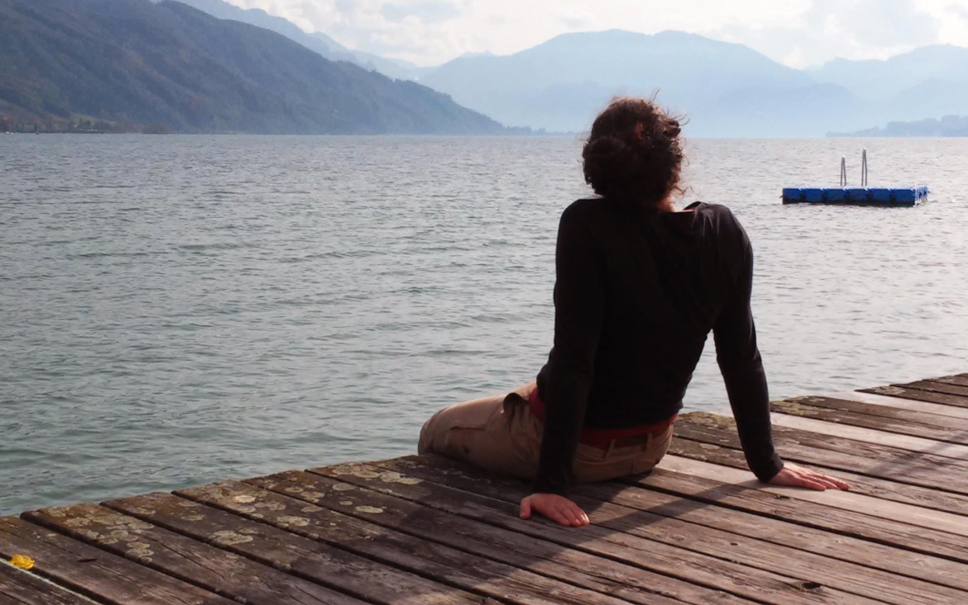 Eine Frau sitzt uns mit dem Rücken zugewandt auf einem Steg mit Blick auf den See