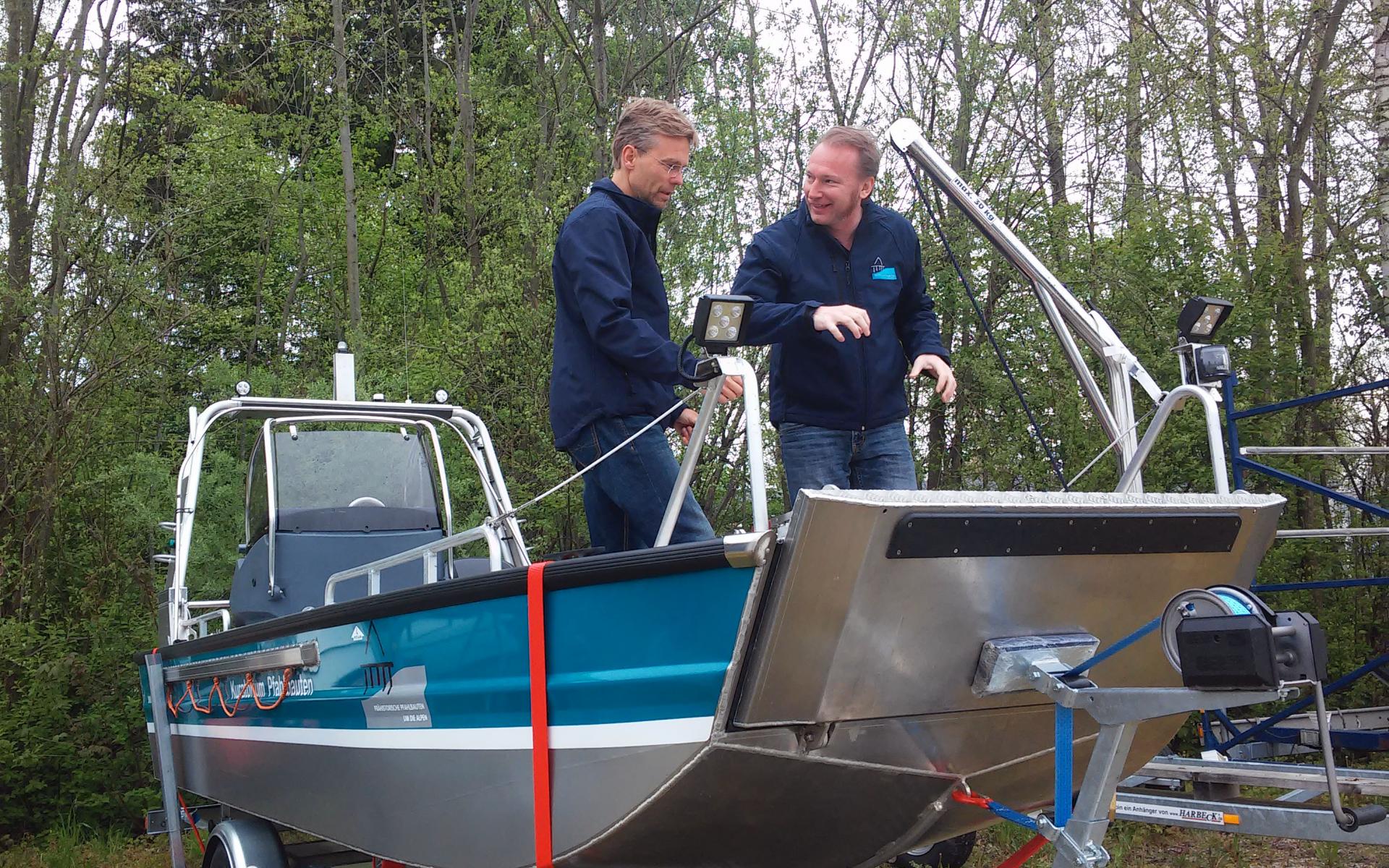 2 Männer stehen auf einem aufgebockten, blau-silbernen Motorboot