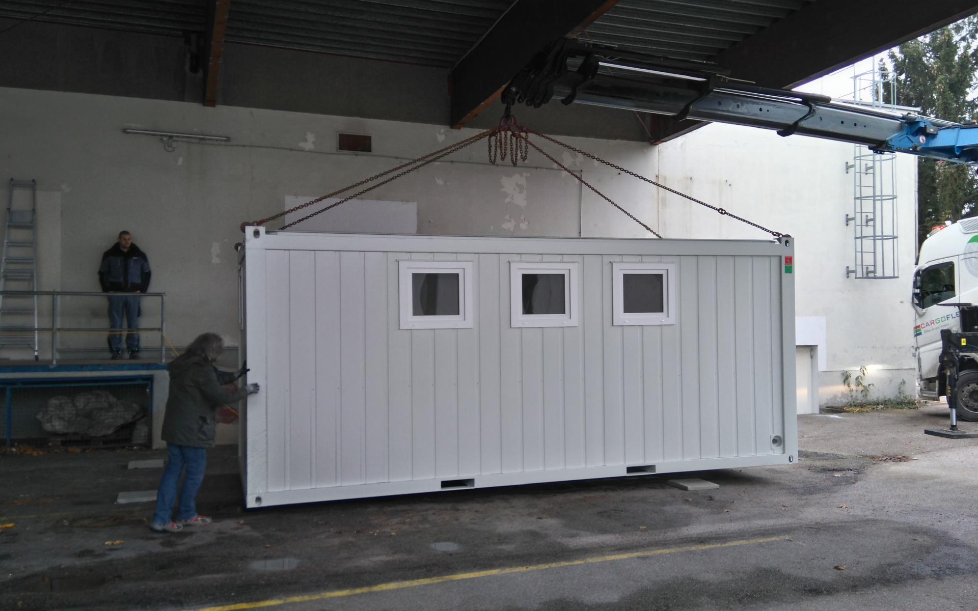 2 Arbeiter und ein Kranwagen heben den weißen Container an seinen Platz