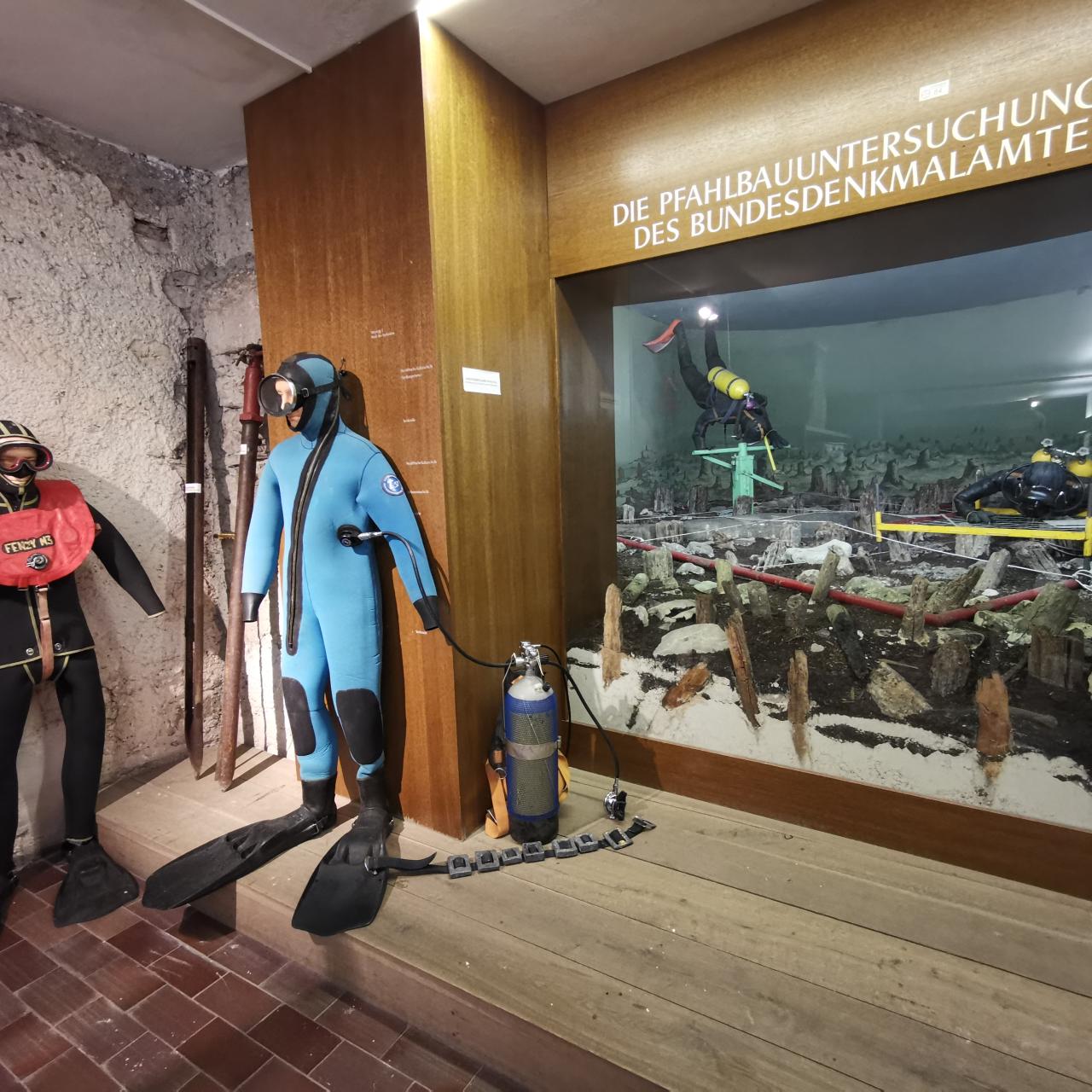 2 Taucheranzüge vor einem lebensgroßen Diorama mit Taucher:Innen