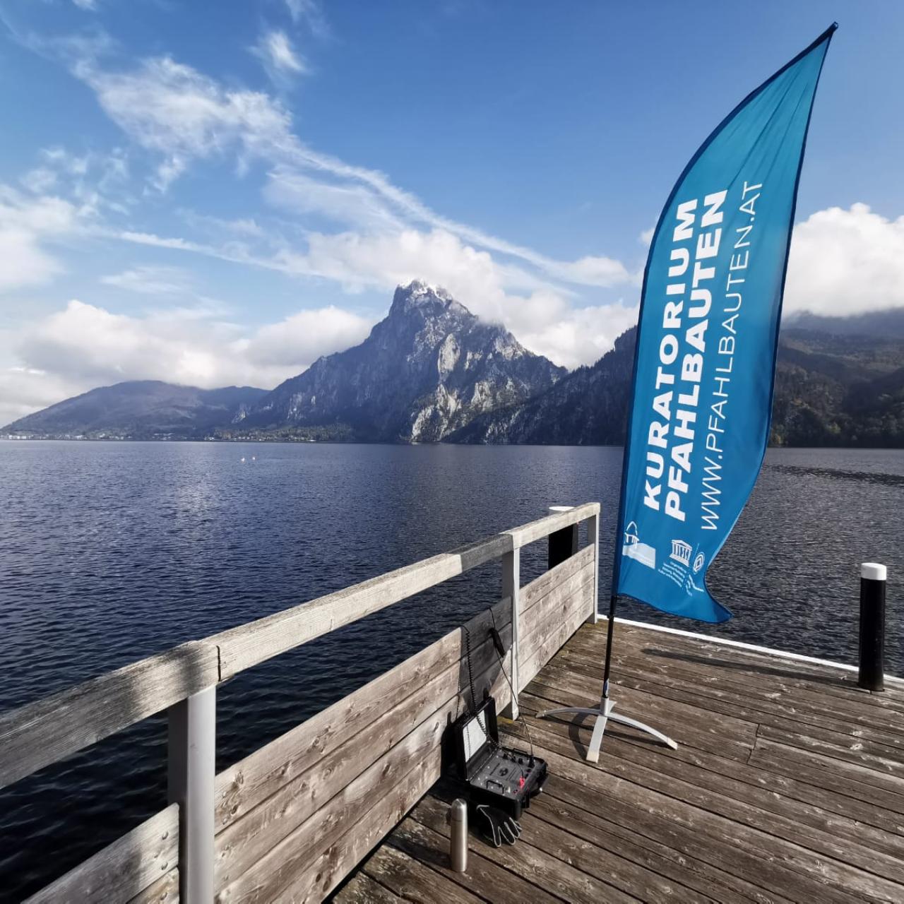 Eine blaue Beachflag des "Kuratiorium Pfahlbauten" auf einem Steg vor dem Traunsee