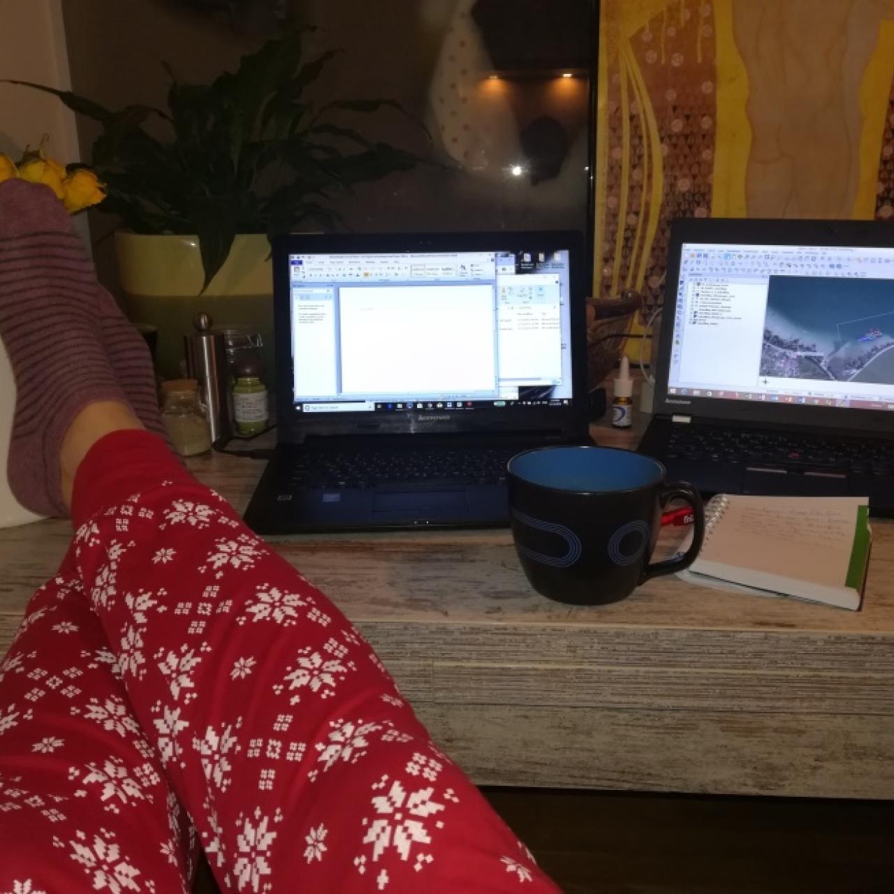 Man sieht Beine in einer roten Pyjama-Hose, die auf einem Schreibtisch liegen. Daneben eine Kaffeetasse und 2 Laptops