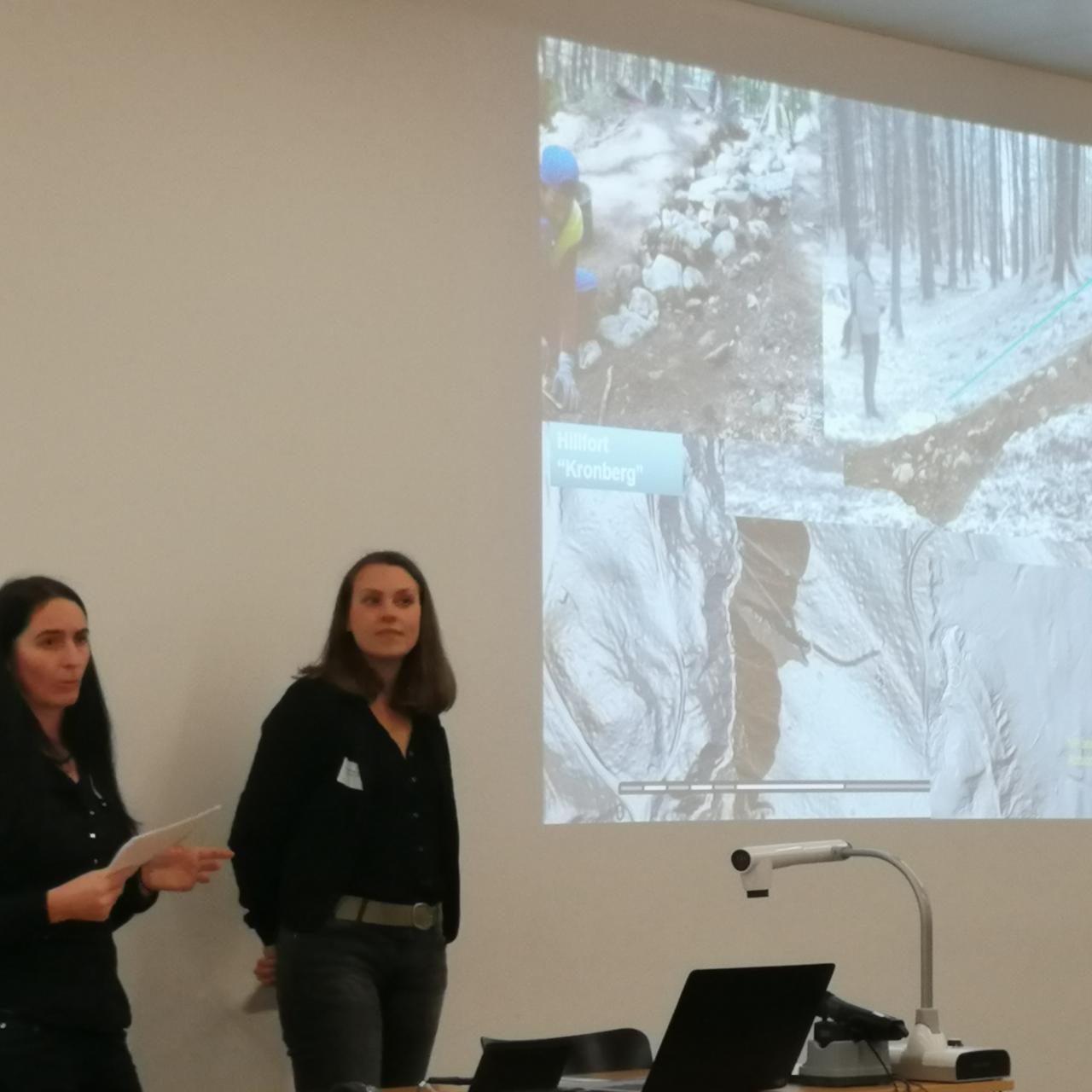 2 Frauen stehen vor einer Leinwand bei einem Vortrag