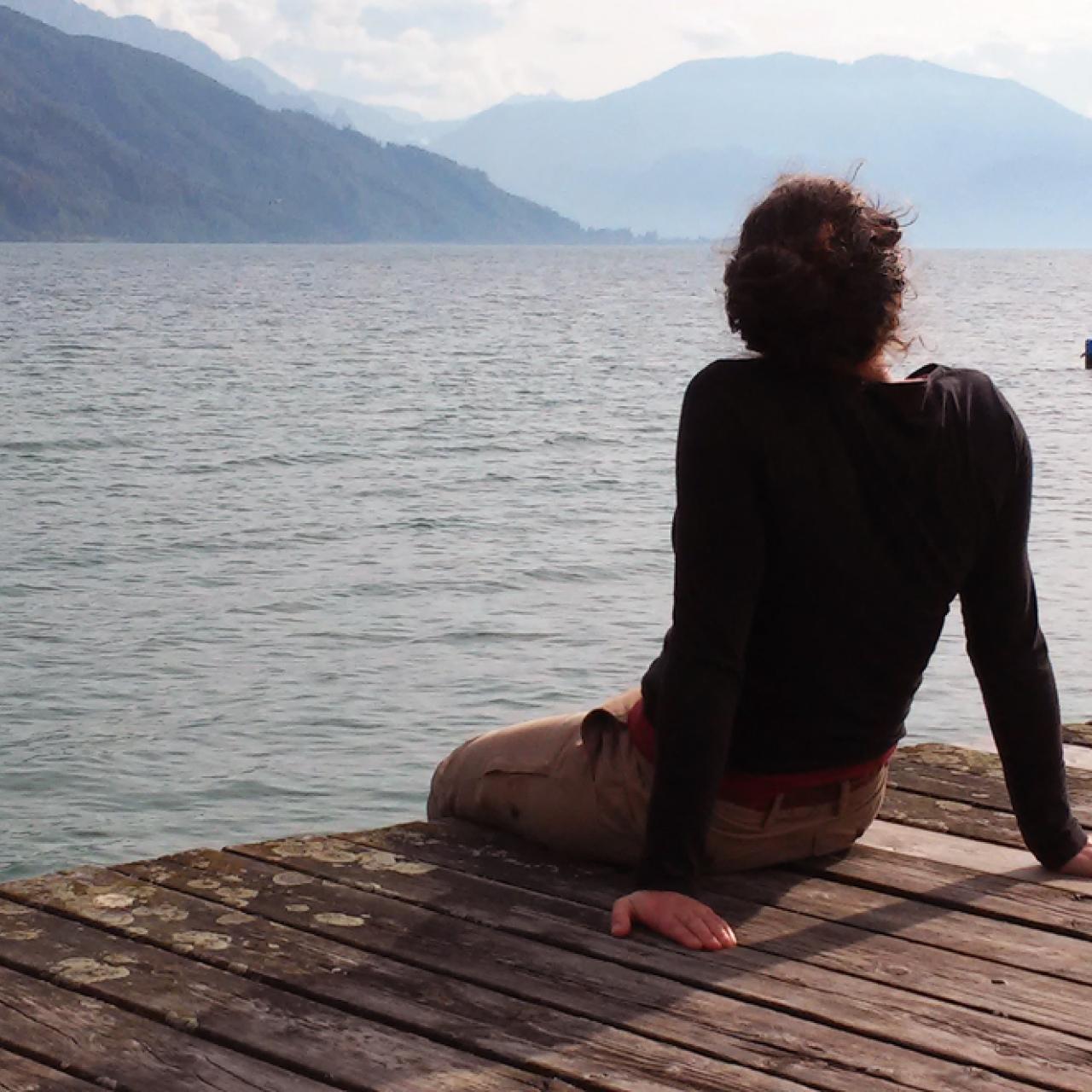 Eine Frau sitzt uns mit dem Rücken zugewandt auf einem Steg mit Blick auf den See