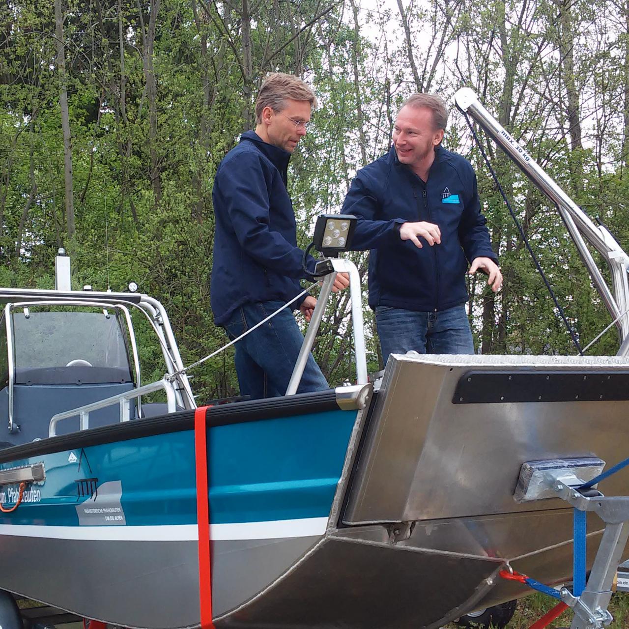 2 Männer stehen auf einem aufgebockten, blau-silbernen Motorboot