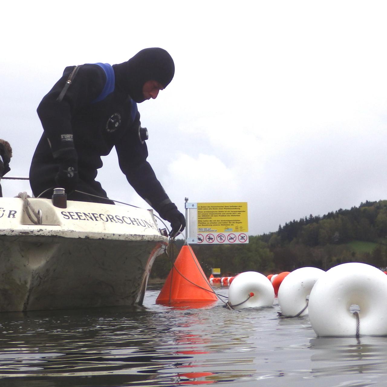 Ein Mann im Tauchanzug an Bord eines Bootes setzt Markierungsbojen in den See